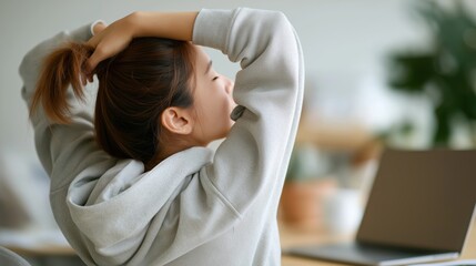 Young woman stretching her arms behind her head while sitting at a desk with a laptop.