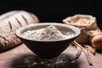 Fresh spelt flour fills a rustic bowl next to bread, eggs, and wheat stalks, ready for baking