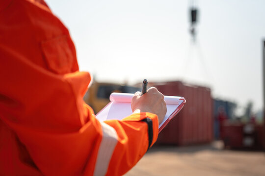 An operation supervisor is writing on paper during perform safety audit at construction work site, with blurred background of heavy lifting operation. Industrial safe work practice concept scene.