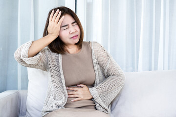 Stressed Asian woman suffering from stomach ache and headache on sofa. Illustrating gut-brain axis connection, digestive issues, and mental stress impact on physical health in a bright living room