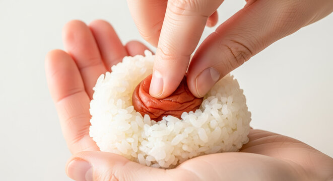 握りたてのおにぎりに梅干しをやさしく押し込む手元のクローズアップ。素朴な素材で作る伝統的な家庭料理の一場面。