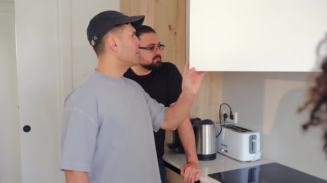 Maintenance technician inspects upper kitchen cabinet while speaking with residents. Indoor apartment repair service, home improvement discussion and property care concept.