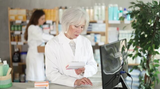 Processing of electronic document by computer. Senior woman employee in labcoat works in pharmacy using computer, remote document flow. Female companion work in background 