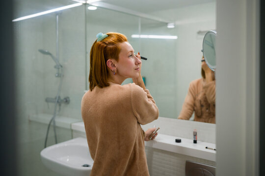 Woman applying makeup in bathroom mirror as part of self care routine