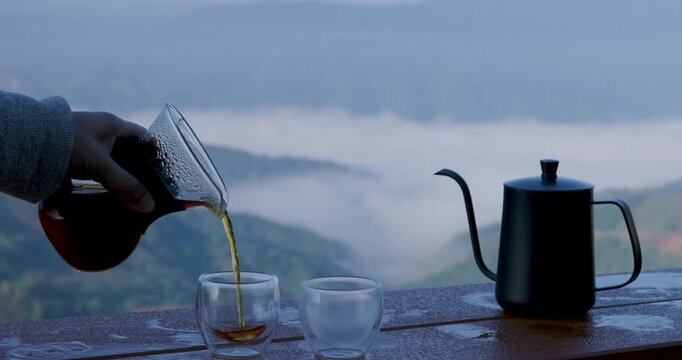 Hand Pouring Hot Drip Coffee Into Glass Cup On Wooden Table With Misty Mountain View Background