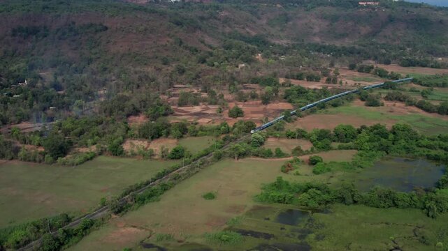 Drone captures blue Indian Railways train approaching along tracks through rural fields, sparse forests, and hilly terrain near Chiplun in Maharashtra.