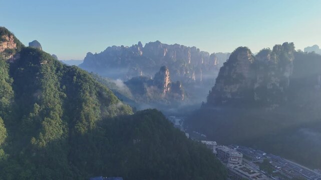 Towering stone spires pierce drifting mist above deep green valleys and cliffs, aerial medium over of Zhangjiajie China village town