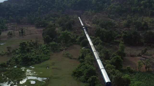Aerial view of long silver Indian Railways train entering and disappearing into hill tunnel, surrounded by lush green forests, fields, and rugged terrain near Chiplun.
