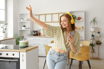 Cool young woman with mobile phone listening to music and dancing in kitchen