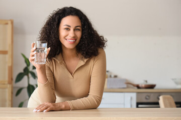 Young African-American woman with glass of water at table in kitchen