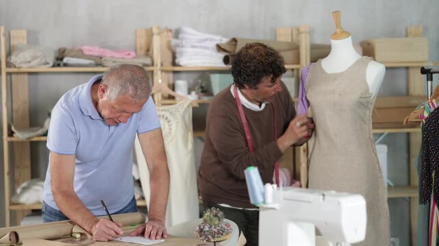 Elderly man tailor drawing pattern on paper adult man assistant sews on mannequin in workshop