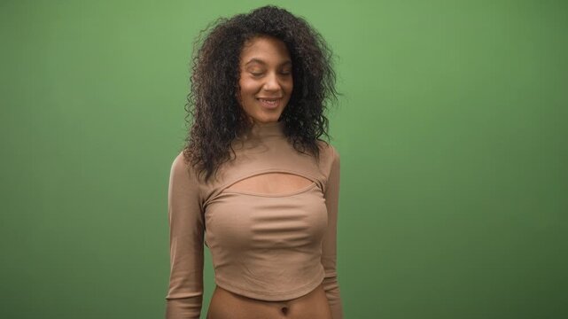 Young woman with curly hair points finger upward with a knowing smile against a green studio backdrop; playful curiosity.