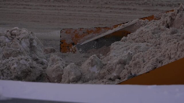 Heavy equipment bulldozer clearing a city road from dirty snow and ice with its front loader bucket, while a municipal service worker with a shovel helps with winter maintenance after a blizzard