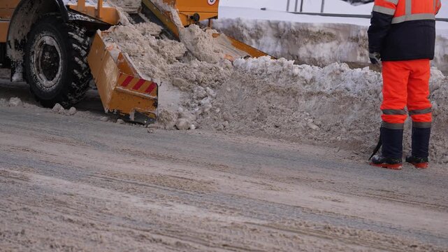 Heavy machinery clearing a city street of dirty snow and ice buildup as a road worker in high-visibility clothing observes, ensuring public safety during cold winter weather conditions