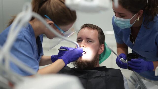 Woman dentist and a nurse carefully drill the tooth of a worried man, they remove caries and prepare the tooth for a filling. Man came to treat his teeth