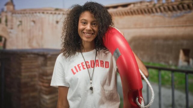 Teenage lifeguard woman holds red lifebuoy on shoulder and smiles wearing whistle necklace in front of historic building; confidence duty.