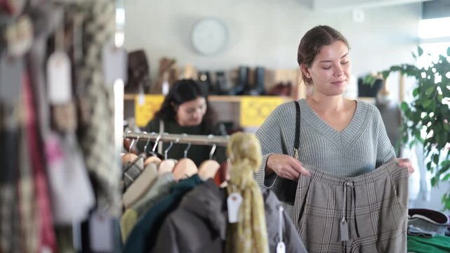 Young American woman in warm clothes chooses woolen beige pants, she stands near the counter with clothes at discounts. Woman makes profitable purchases in a store. High quality 4k footage