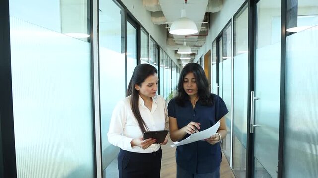Two young indian female boss or manager with gen z girl discuss discussing work using digital tablet and paper documents while standing in a modern office corridor. Teamwork and communication.