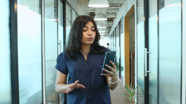 Young indian business gen z girl video call on smart phone while walking in modern office discuss work or project. Employee Online distance meeting. Job opportunities.