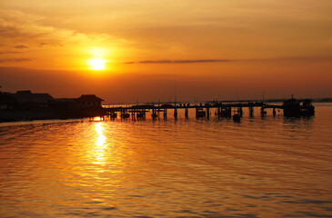 Obraz premium The sunset at the sea village, the sky glows in warm shades of orange and gold as the sun sinks low on the horizon, its light shimmering. A wooden pier stretches outward, with small boats alongside.
