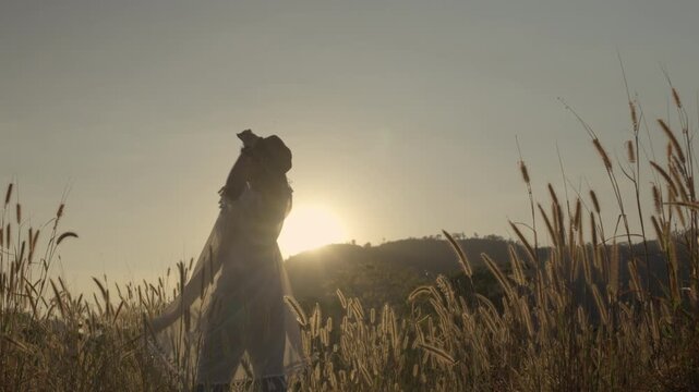 Asian woman in boho style with feather headband walking and spinning on grassy hill at sunset with mountain background and lens flare.