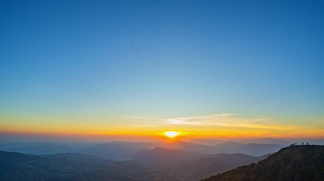 In a smooth flow of changing light, the sun emerges beyond towering peaks, transforming the sky into a glowing canvas. The time lapse reveals the tranquil charm of dawn across the vast mountain range.