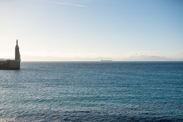 Fototapeta premium Wide ocean view with lighthouse statue on pier distant cargo ship and hazy mountains under soft sunlight creating tranquil maritime landscape with open water and horizon