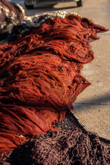 Fototapeta premium Red fishing nets drying on concrete harbor pier in warm sunlight, showing layered mesh textures, traditional maritime equipment, coastal fishing industry atmosphere, and weathered materials