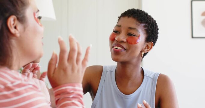 Diverse female friends reacting to hand gestures, sharing skincare at home with under-eye patches