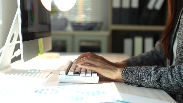 Professional woman typing on a modern computer keyboard while working on business data charts and graphs in a sunlit office, showcasing productivity, technology, and financial analysis