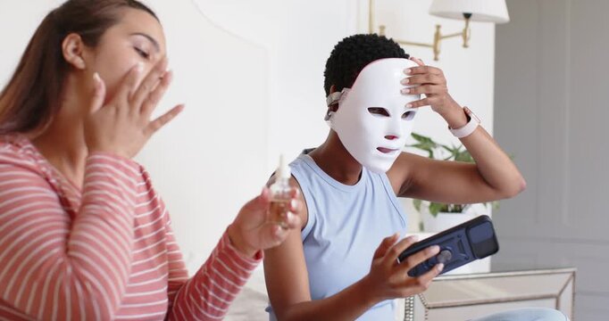 Diverse female friends on bed left using serum, right fitting LED mask, tapping remote for skincare