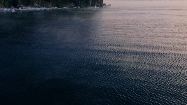 Low-angle aerial approach over the rippling water of Lake Superior, revealing Split Rock Lighthouse through thick morning sea smoke.