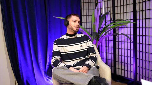 Young man wearing headphones speaking and gesturing during a live interview or podcast recording in a modern, colorful content creation studio with purple and blue lights