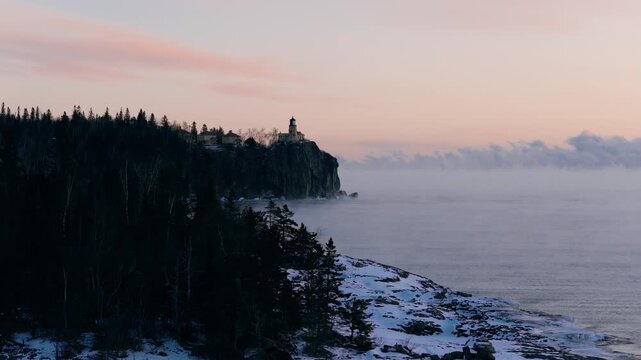 Aerial view looking through pine trees toward Split Rock Lighthouse as it stands guard over a misty winter lake.