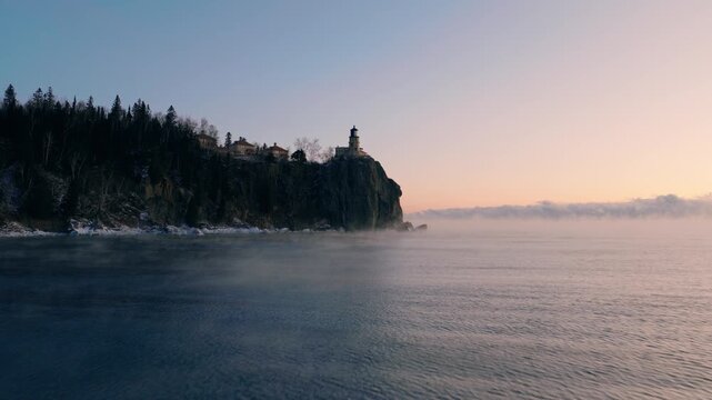 A steady low-altitude aerial shot moving across the rippling waters of Lake Superior toward Split Rock Lighthouse during a misty winter dawn.