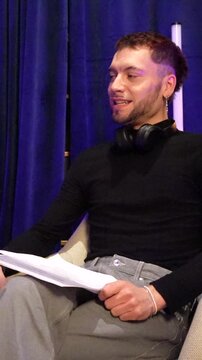 Confident young man with headphones around his neck speaking and gesturing while holding a script during a podcast interview, sharing his personal story in a professional studio setting