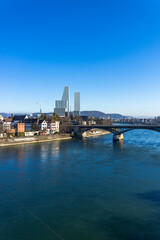 Vertical panorama of the Rhine river in Basel, Switzerland, featuring the Wettstein Bridge and the modern Roche Towers under a clear blue sky.