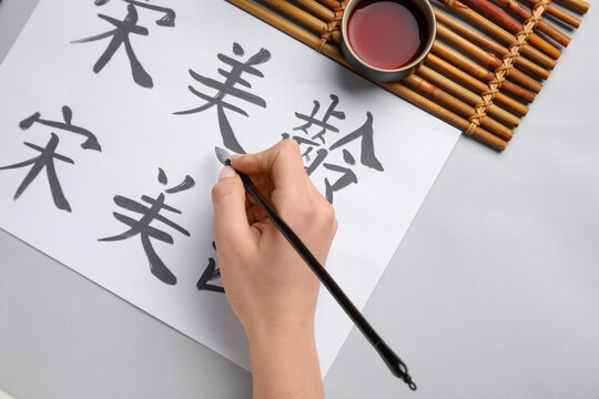 Female hand with Asian hieroglyphs, brush and cup of tea on light background, closeup. International Haiku Poetry Day