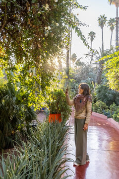 Woman takes a photo in a lush garden with palm trees and aloe plants, sunlight filtering through the trees. Jardin Majorelle,Marrakech,Morocco
