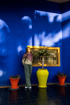 A woman stands in front of a bright blue wall with potted plants and a yellow window frame. Jardin Majorelle,Marrakech,Morocco