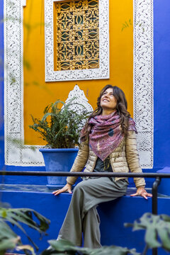 A woman with long brown hair, wearing a scarf and jacket, sits on a blue ledge, looking up at a yellow wall with white trim and a decorative window. Jardin Majorelle,Marrakech,Morocco