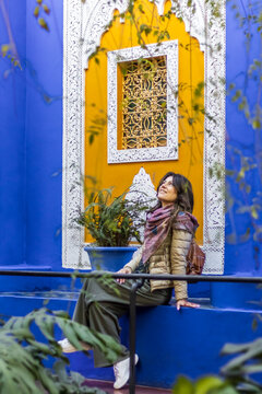 A woman sits on a blue ledge, looking up at a yellow wall with white trim and a decorative window. Jardin Majorelle,Marrakech,Morocco