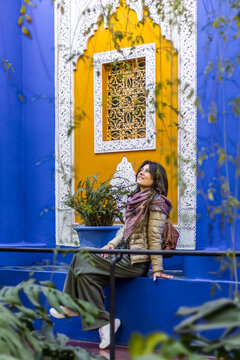 A woman sits on a blue ledge, gazing up at a yellow wall with a white-framed window. Lush greenery surrounds her. Jardin Majorelle,Marrakech,Morocco