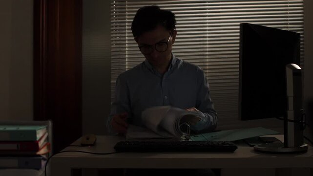 Young man in glasses working late at a dim office desk, focused on paperwork and computer by window blinds, studying documents under soft evening light for a deadline