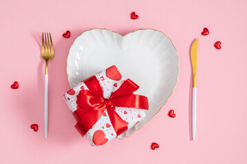 Valentine's Day romantic table setting: White heart-shaped plate with gold rim, gift box with red ribbon, golden fork and knife on pink background. Flat lay concept for wedding or anniversary.