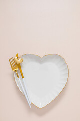 Elegant table setting: a white ceramic heart-shaped plate with a gold rim, a gold fork, and a knife on a pastel background. Concept for a romantic dinner, wedding, or Valentine's Day. Top view, flat l