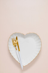 Romantic table setting: a white ceramic heart-shaped plate with a gold rim, elegant gold-tipped flatware, and a small gold heart on a light background. Valentine's Day, wedding. Top view, flat lay.