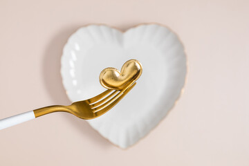 Golden heart on an elegant golden fork against a white ceramic heart-shaped plate. Concept for a romantic dinner, Valentine's Day, wedding, or special occasion. Top view