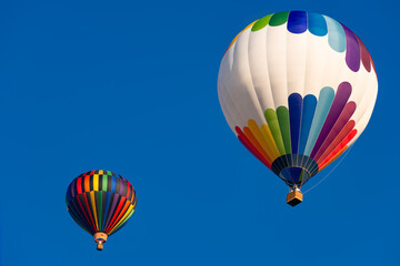 Obraz premium Two colorful hot air balloons with brightly colored envelopes and balloonists in the baskets hanging below on a sunny day in the Sauerland region (Germany), set against a cloudless blue sky. 