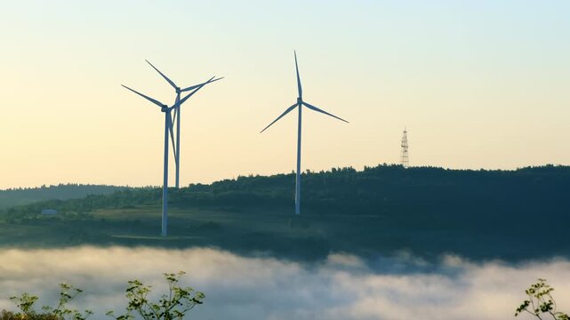 Wind generators operate while haze flows in hill bottom against blue sky. Windmills produce sustainable electric power at highland slow motion